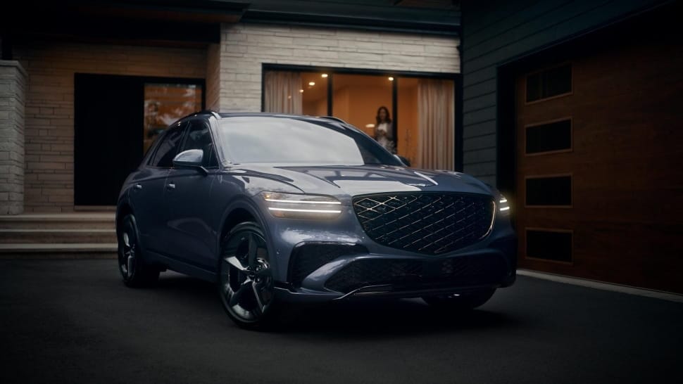 A woman looks out from the window in her house towards a Genesis GV70 in her driveway.