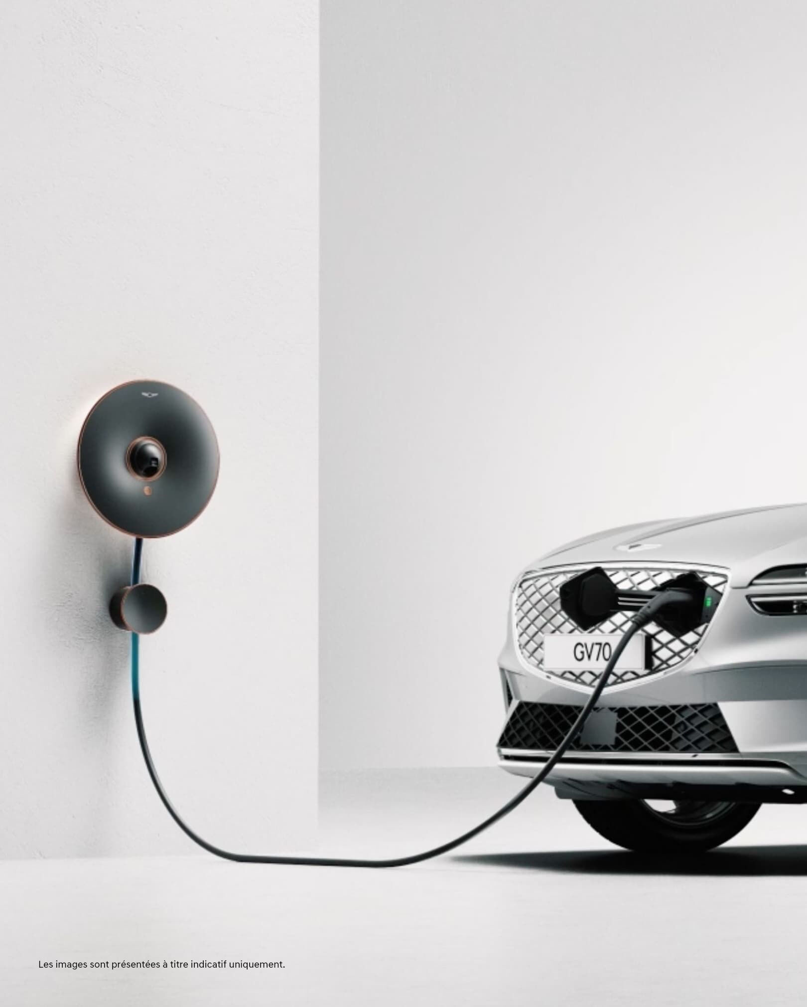 Le GV70 Électrifié est en cours de chargement dans une pièce blanche. Le chargeur connecte le mur au port de recharge dissimulé de la calandre G-Matrix.