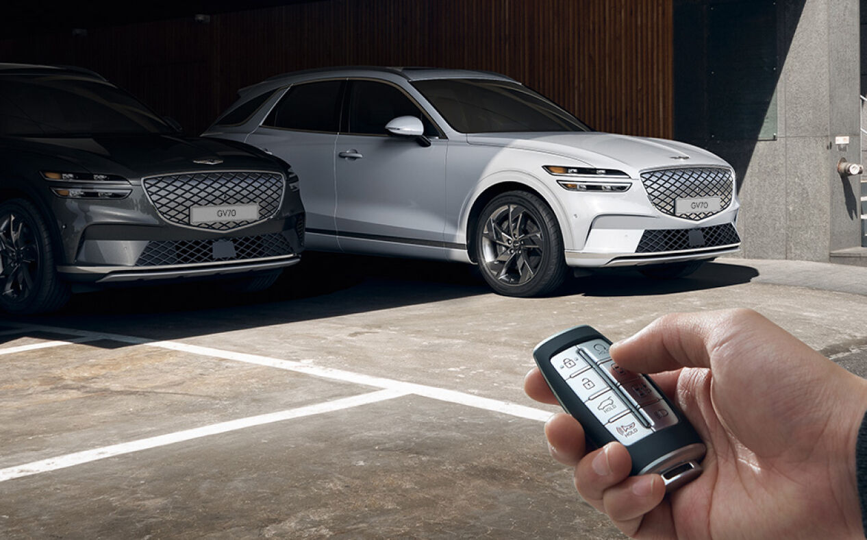 The Electrified GV70 is seen in the distance parked in a parking lot as a person holds a key and presses the Remote Smart Parking Assist button.