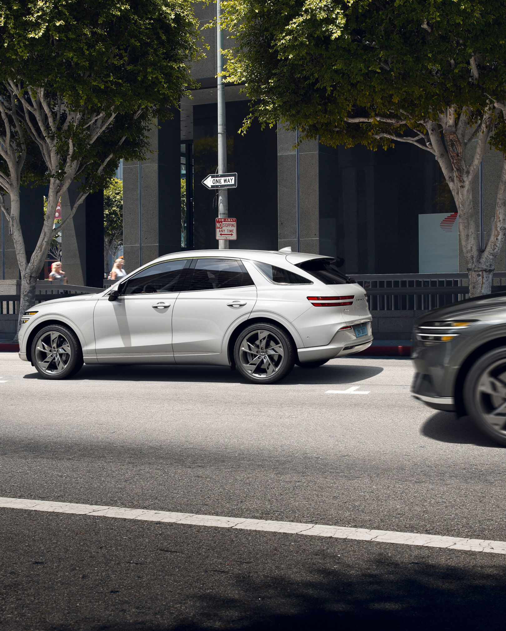 The Electrified GV70 is parked on the street as another car passes by from behind.