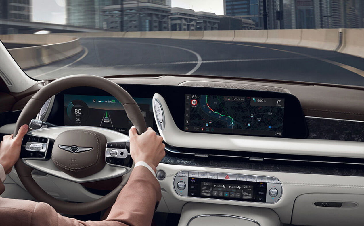 The driver holds the steering wheel and navigation is visible on the infotainment display in Glacier White interior.