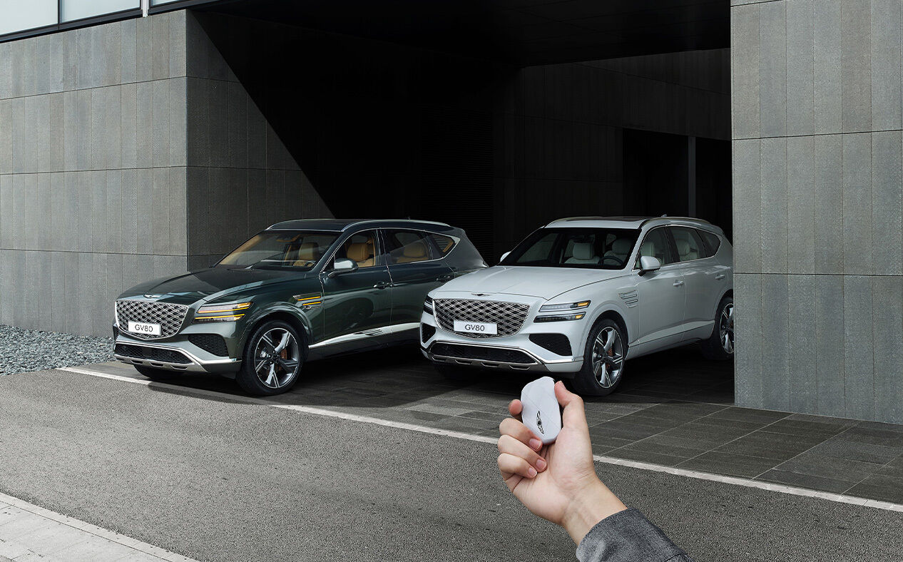 A Vik Black GV80 and a Uyuni White GV80 are parked, and the driver is operating the smart key outside the vehicle.