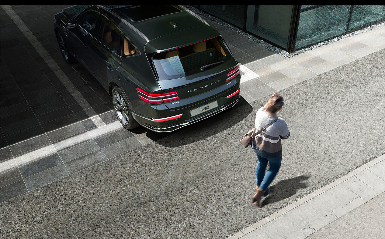 Storr Green GV80 backing out of a parking lot, and a woman is passing behind the vehicle.