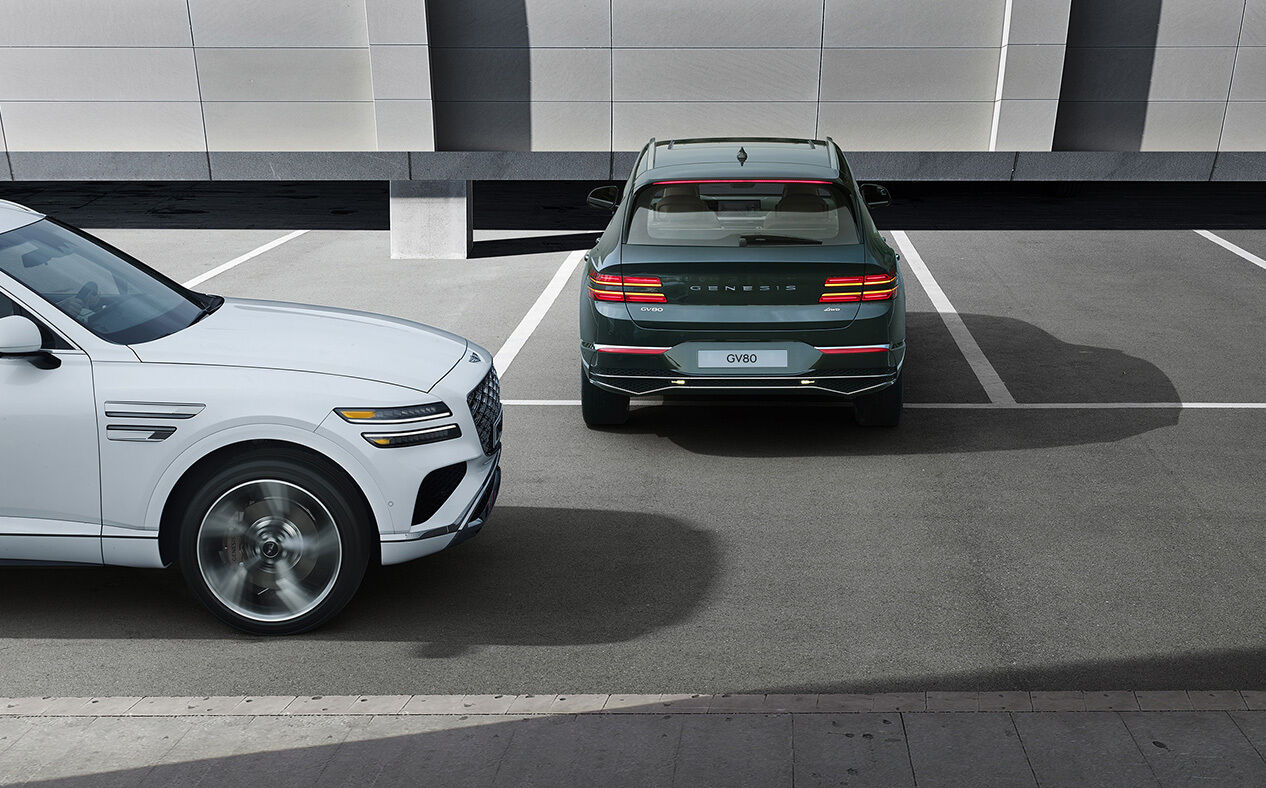  Storr Green GV80 is backing out of a parking spot, and a Uyuni White GV80 passes behind the vehicle on the left.