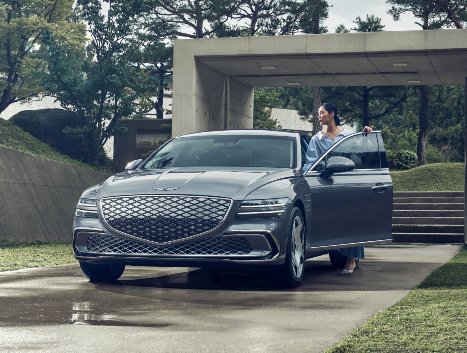 콘크리트 벽과 나무가 어우러진 실외 공간에서 Electrified G80 차량의 운전석에 탑승 중인 여성이 보입니다.