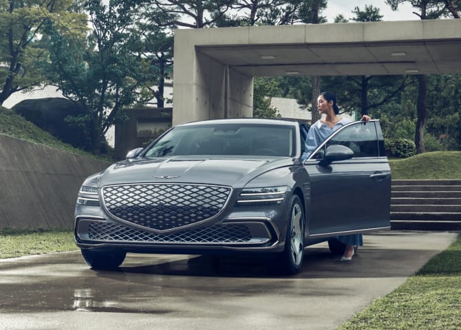 콘크리트 벽과 나무가 어우러진 실외 공간에서 Electrified G80 차량의 운전석에 탑승 중인 여성이 보입니다.