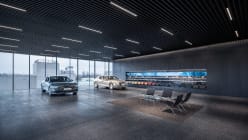 On the left side, various fabric pieces representing Genesis brand colors are displayed above a dark-toned wall cabinet. In the center, a Genesis display vehicle is visible, placed behind a carpeted area where chairs and tables are arranged.