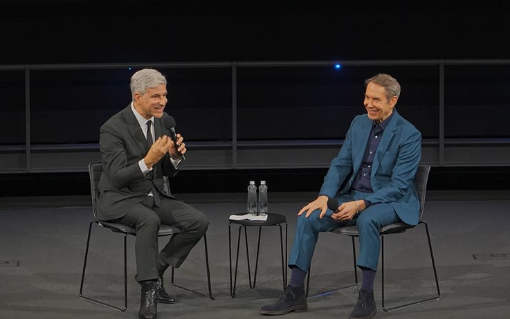 Two men seated on a stage having a conversation; the man on the left speaks into a microphone.