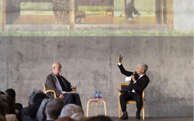 Two men in conversation on a stage; the man on the right raises his hand while speaking, with a large image projected behind them and an audience seated in front.