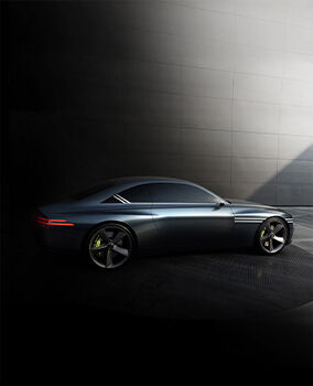 A side view of the Genesis X Concept vehicle parked in a darkened interior, illuminated by natural light streaming in from the upper right of the room. This light reflects off the bodywork, emphasizing the metallic color of the car's surface.