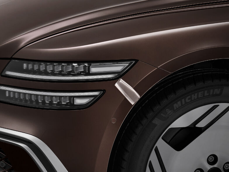 A zoomed-in view of the headlamps and the area near the front wheels of a Brooklyn Brown colored Electrified G80. It gives the impression that some of the vehicle's protective film applied to the body is peeling off.