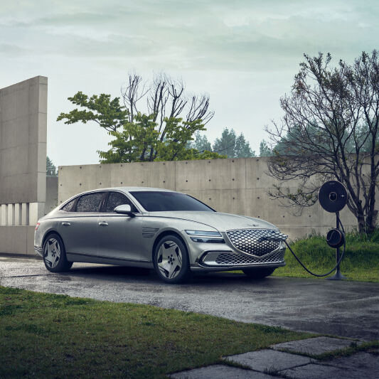 An Electrified G80 is charging with the Genesis EV Home Charger in an outdoor area setting with an exposed concrete wall. The view is from the left side, with the vehicle connected to the nozzle of the charger.