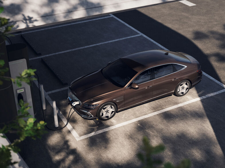 An Electrified G80 is charging in an outdoor electric vehicle charging station. The shadow of a tree is casting over a car parked in the parking line.