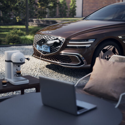 On a sunny day, an Electrified G80 is charging in a graveled garden area. A laptop on a coffee table, a small coffee machine, and more are visible in front of the car, creating a relaxed atmosphere.