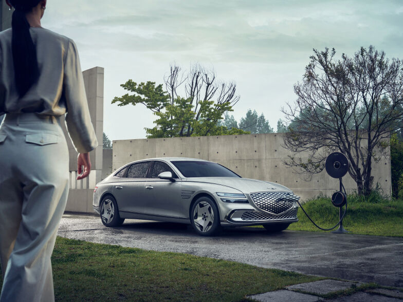 An Electrified G80 is charging with a Genesis EV Home Charger in an outdoor area setting with an exposed concrete wall. A woman is seen in the rear view approaching the vehicle.