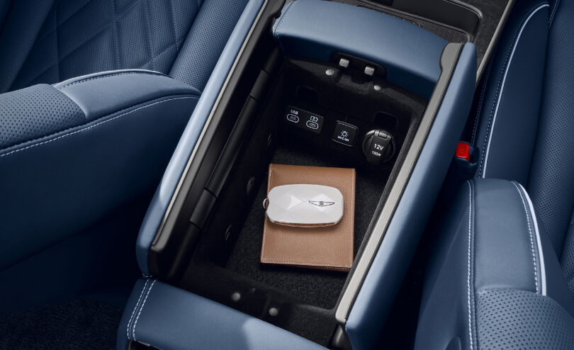 You see two charging outlets, a UV-C ON button, and a 12V/180W power socket in a storage bin in the front seat console armrest. The bin contains a smart key with the white Genesis logo and a light brown wallet.