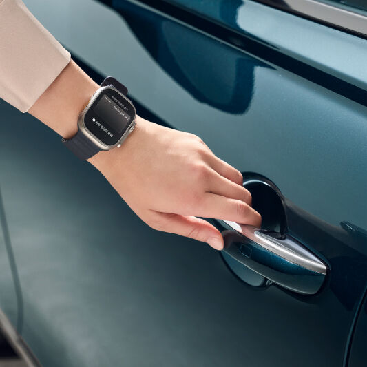 A hand wearing a smartwatch is close to a car door, attempting to remotely operate it. 