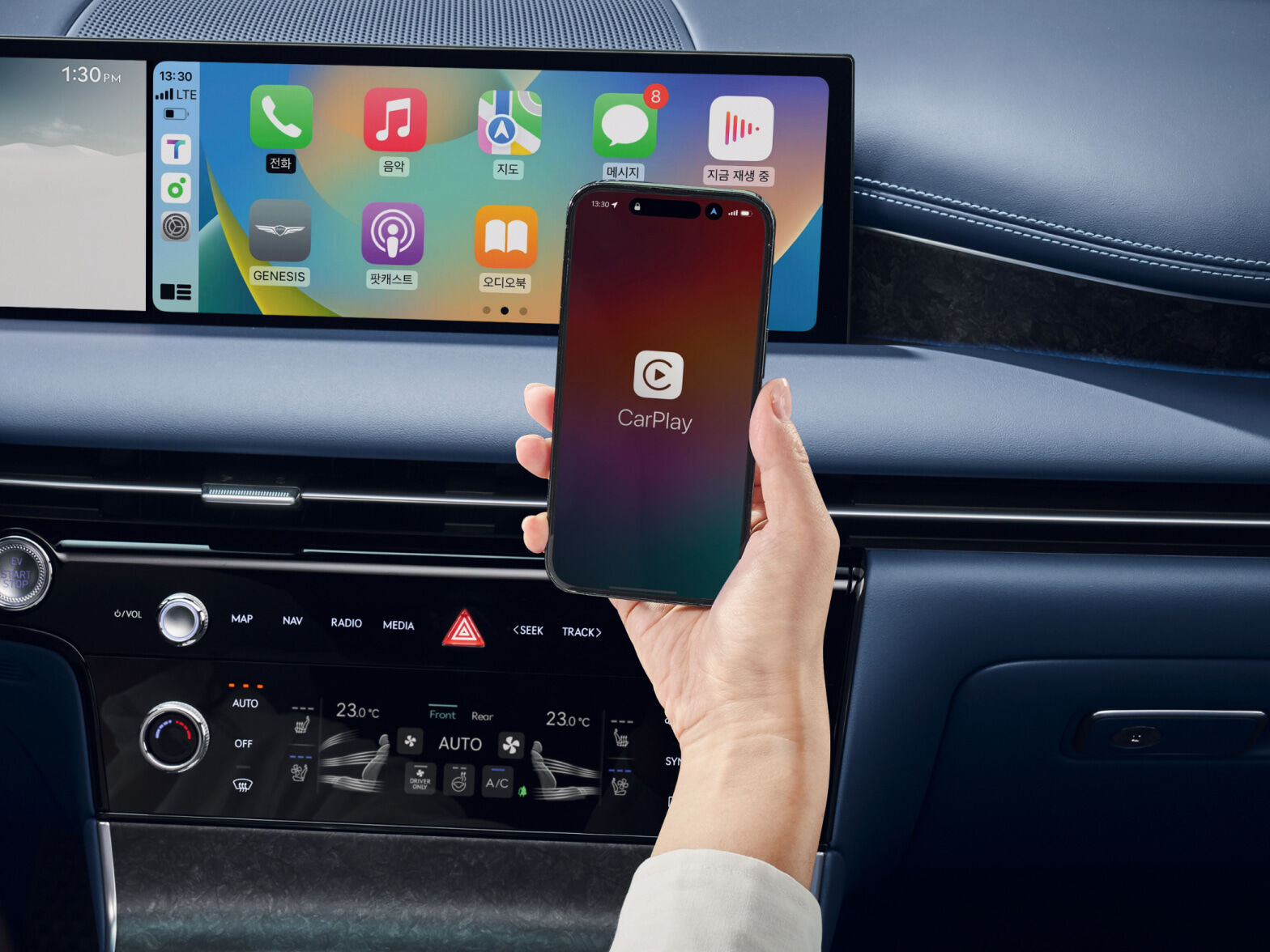 Behind the panoramic display in the front of the front seat, a hand holding a smartphone is visible. The smartphone screen shows the message “CarPlay”.