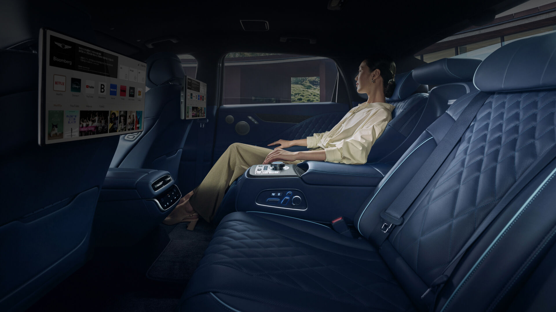 A young woman is shown sitting in the back of a vehicle, resting her arms on the centrally located armrest. The side view shows a variety of content on the screen of the smart infotainment system placed in the headrest of the front seat.