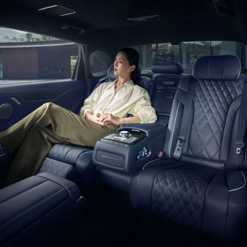 A young woman is shown sitting in the back of a car, resting her arm on the centrally placed armrest. The view is seen from the front seat, giving the impression of a spacious back seat.