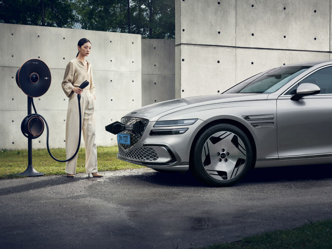An Electrified G80 is parked in an outdoor space, against an exposed concrete wall. A young woman is standing in front of the vehicle with a Genesis EV Home Charger in her hand. The charging door, located in the front crest grille of the vehicle, is open.