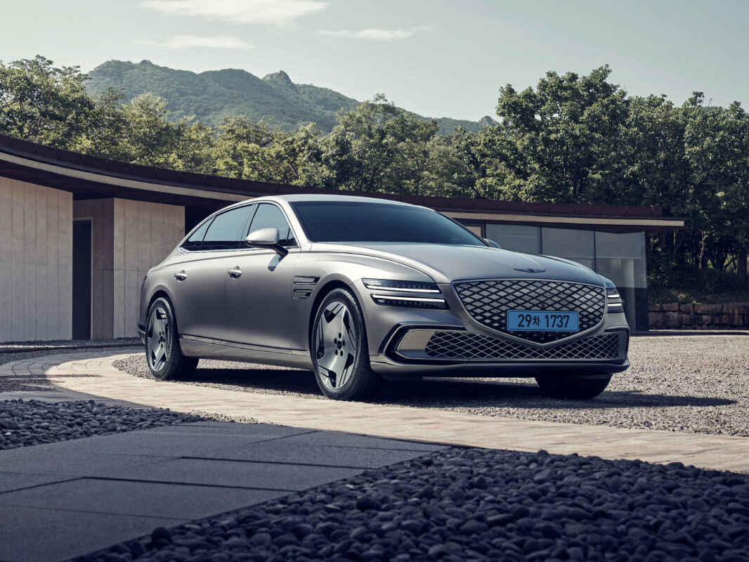 An Electrified G80 in Geneva Silver color is parked in front of a wide-spread building offering a clear view of the front and sides. Behind it lies a vast expanse of buildings, mountains, and sky.