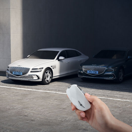 An electrified G80 is moving forward, leaving a shaded parking space, and in front of it you see a hand grasping the car keys.