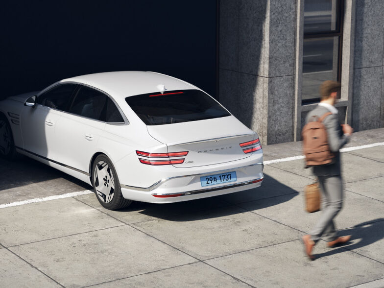 A glossy Uyuni White Electrified G80 trying to reverse and a man passing in front of it.