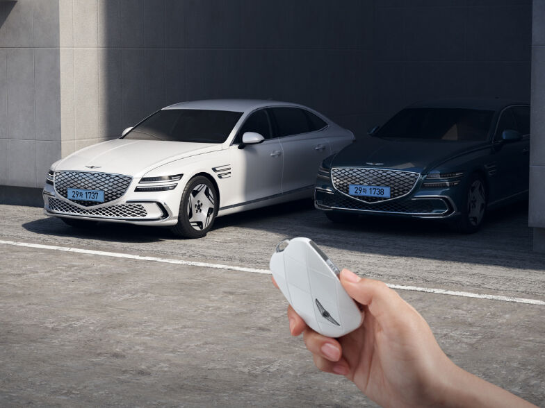 An electrified G80 is moving forward, leaving a shaded parking space, and in front of it you see a hand grasping the car keys.