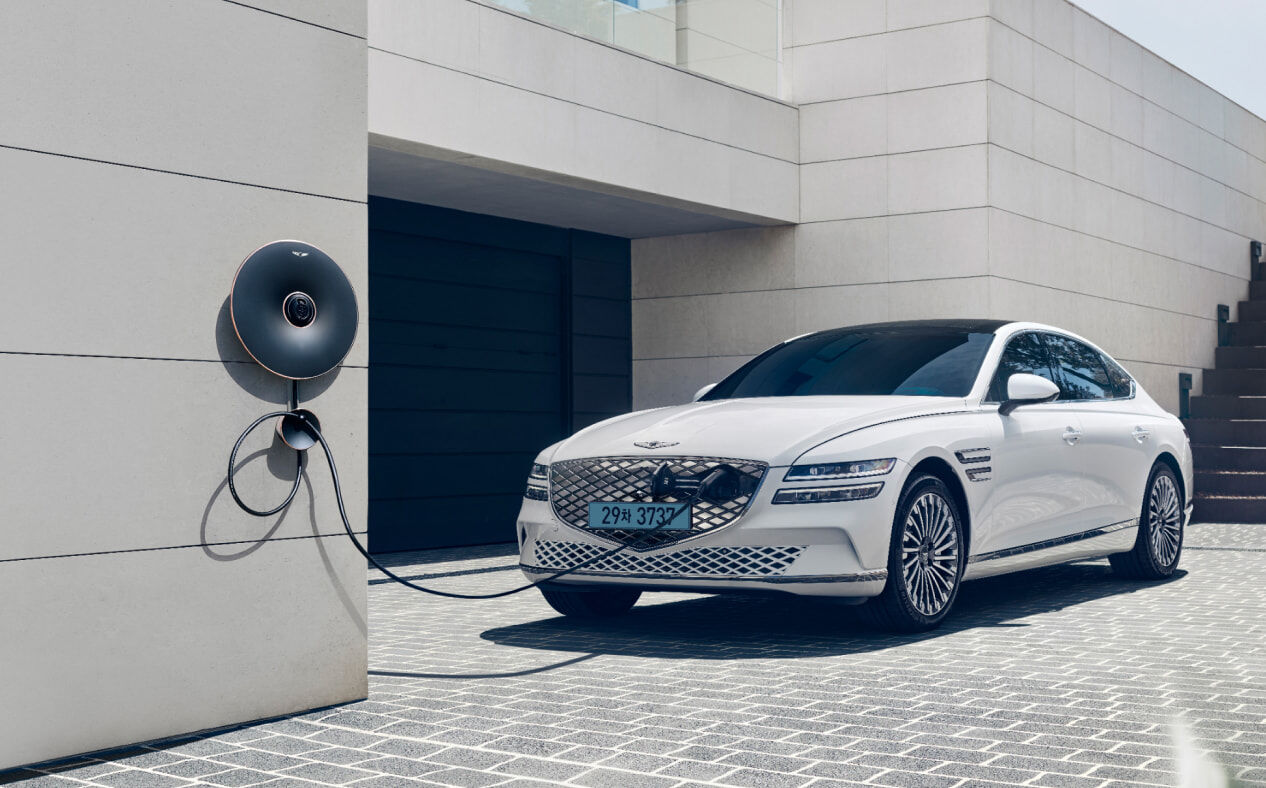 Electrified G80 vehicle in glossy Uyuni White being charged through a home charger. A cable is connected to the hidden parting front charging port to show the vehicle being charged.