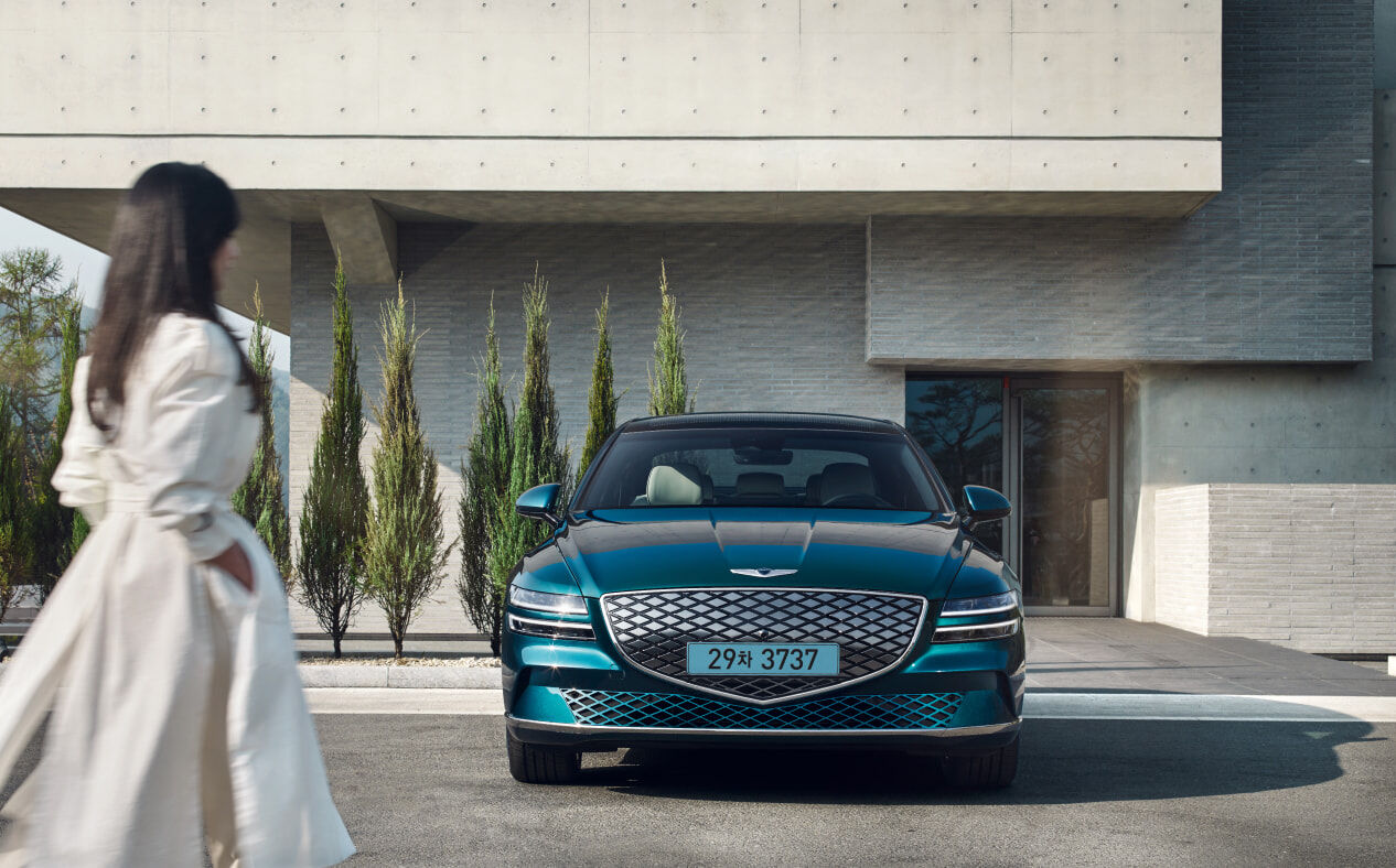 A woman walking towards a parked Electrified G80 in glossy Matira Blue.