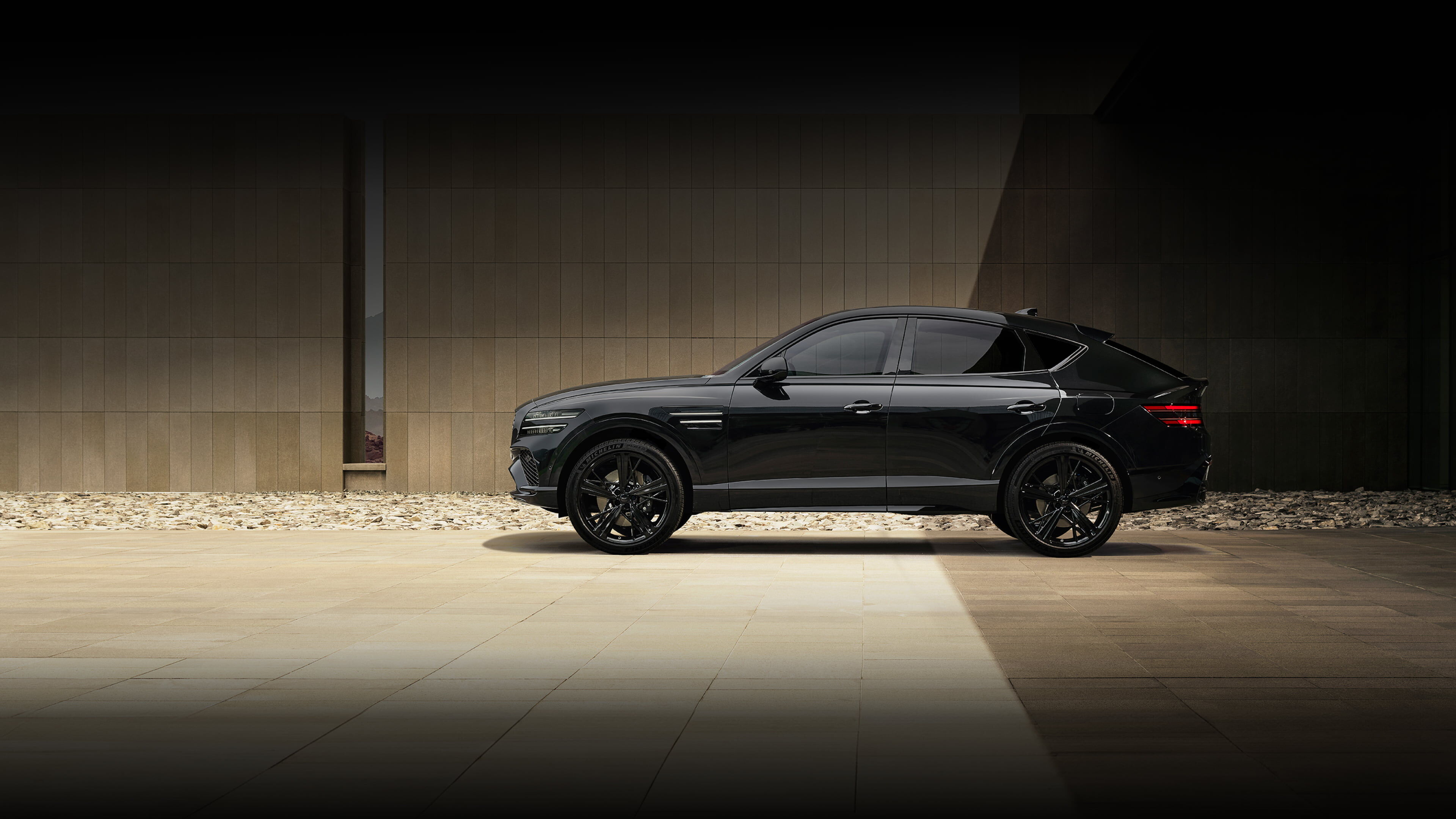 The side of the GV80 Coupe Black is visible in front of a wall made of brown tiles. The rear of the vehicle appears darker due to the shadow cast by the building.