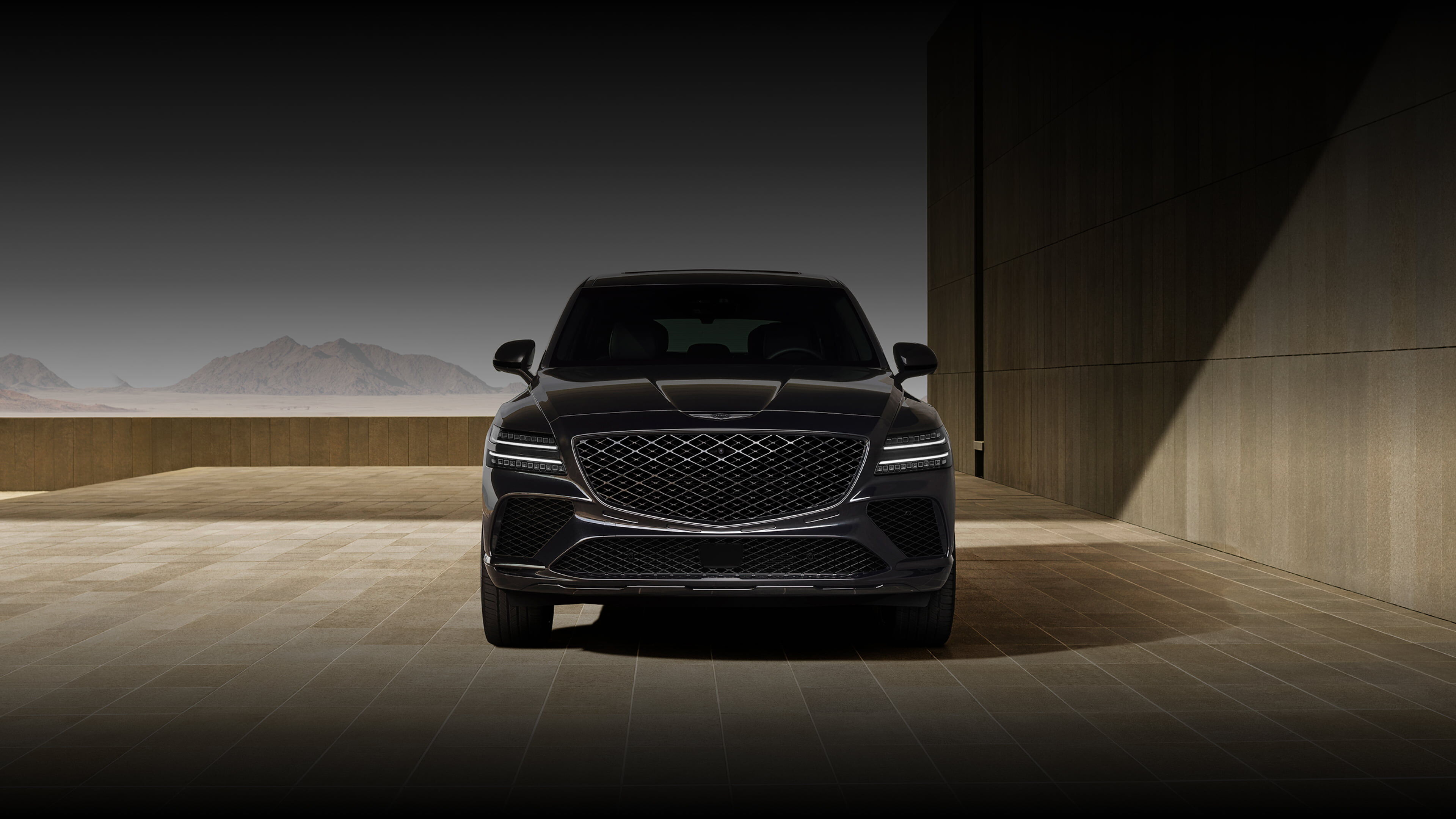 To the right, a wall made of earthy-colored tiles frames the view of the front of the GV80 Coupe Black. Behind the vehicle, a low wall of brown tiles stretches out, with green leaves and the silhouette of a mountain visible beyond