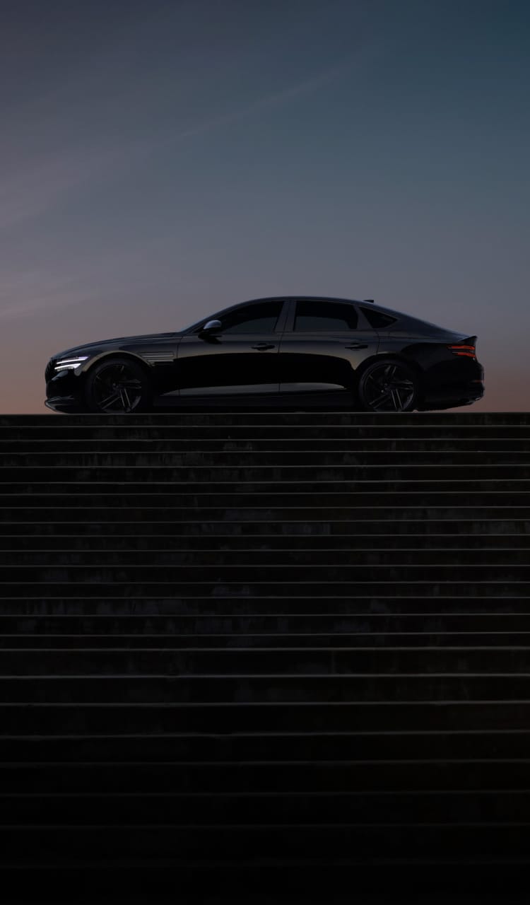 계단 위의 공터에 G80 Black이 좌측면을 보이며 서 있습니다. 차량의 부드러운 측면 라인이 강조되어 보입니다. 그 뒤로 저녁 무렵의 노을에 물든 보랏빛의 하늘이 펼쳐져 있습니다.