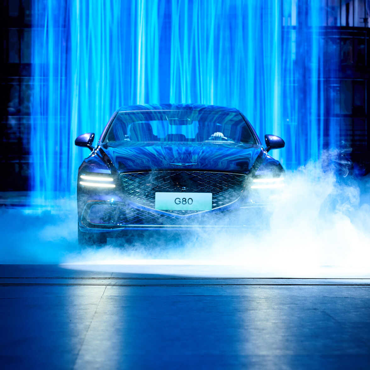 Blue-colored Genesis Electrified G80 Magma Concept's headlights are on, covered in a blue backdrop and fog.