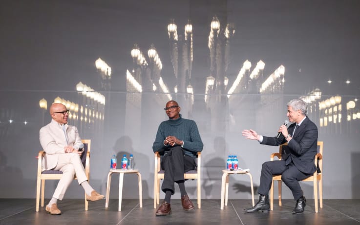 Three men seated side by side on a stage during a panel discussion, with bright lights glowing behind them.