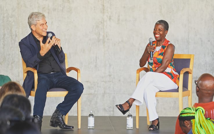 A man and a woman seated on a stage in conversation; the man on the left speaks into a microphone.