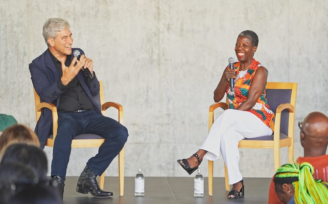 A man and a woman seated on a stage in conversation; the man on the left speaks into a microphone.