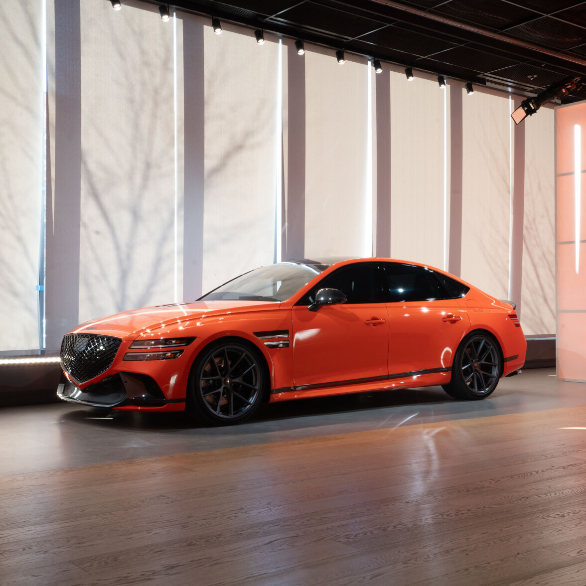 Genesis G80 Magma Special is parked indoors, facing left with its side profile showing. Behind the vehicle are blinds drawn to prevent sunlight. 