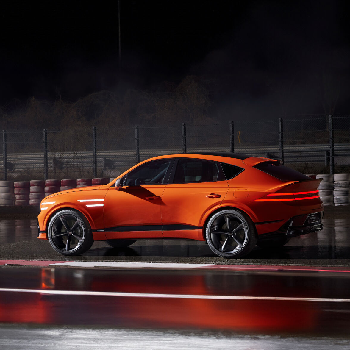 The GV80 Coupe Concept is parked on a racetrack, showing its side. The front and rear lights are brightly lit.