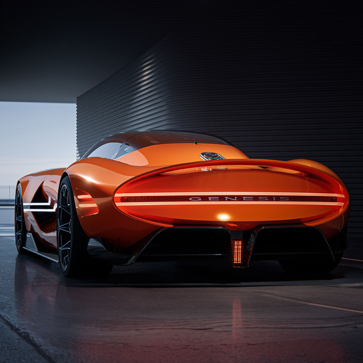 The Genesis X Gran Berlinetta Concept stands poised in front of a dark panel wall, illuminated by white overhead lights. Its side profile and lit headlamps reflect elegantly off the sleek, glossy floor.