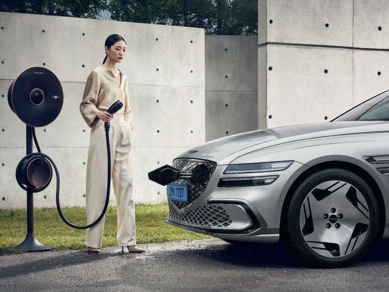 An Electrified G80 is parked in an outdoor space, against an exposed concrete wall. A young woman is standing in front of the vehicle with a Genesis EV Home Charger in her hand. The charging door, located in the front crest grille of the vehicle, is open.