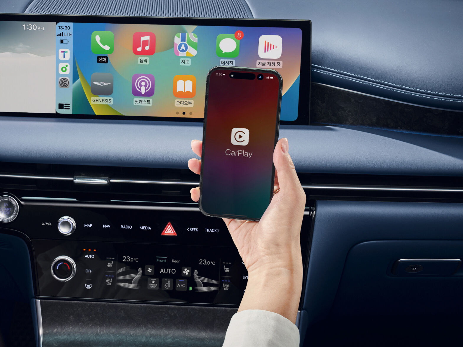 Behind the panoramic display in the front of the front seat, a hand holding a smartphone is visible. The smartphone screen shows the message “CarPlay”.