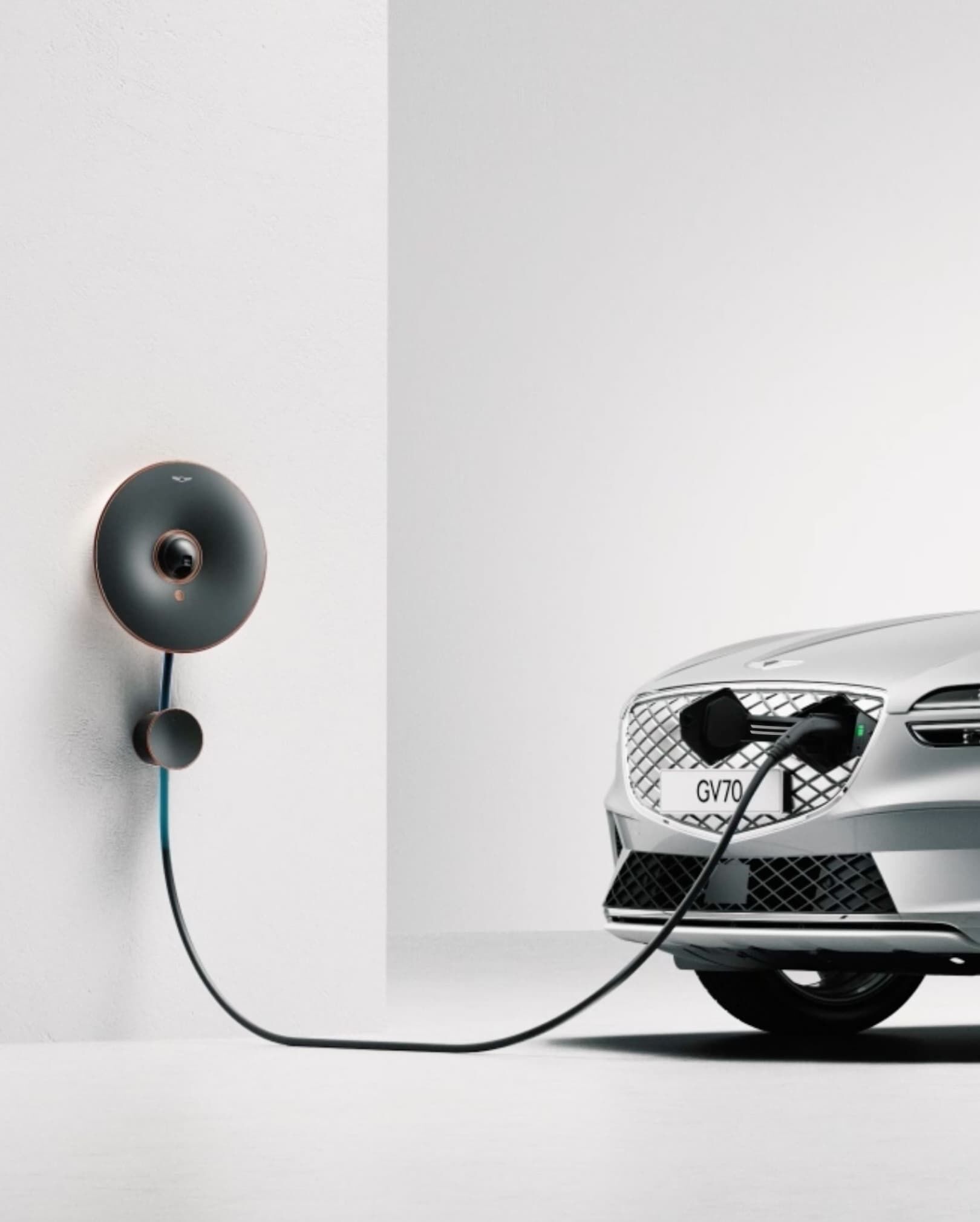 Electrified GV70 is charging in a white room. The charger connects from the wall to the G-Matrix Patterned Grille’s Hidden Charging Door.