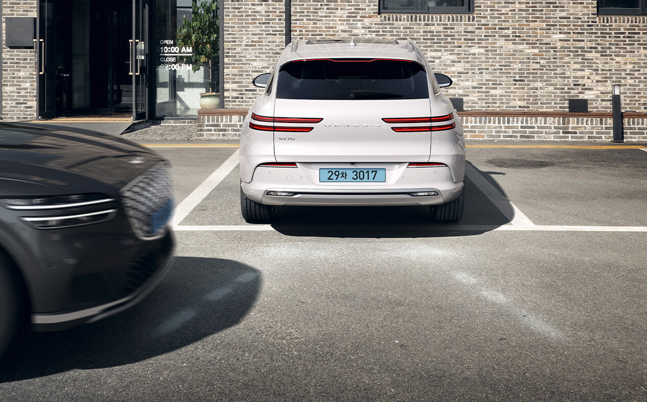 The Electrified GV70 reverses in the direction of a passing car with white lines depicting Reverse Parking Collision Avoidance Assist.
