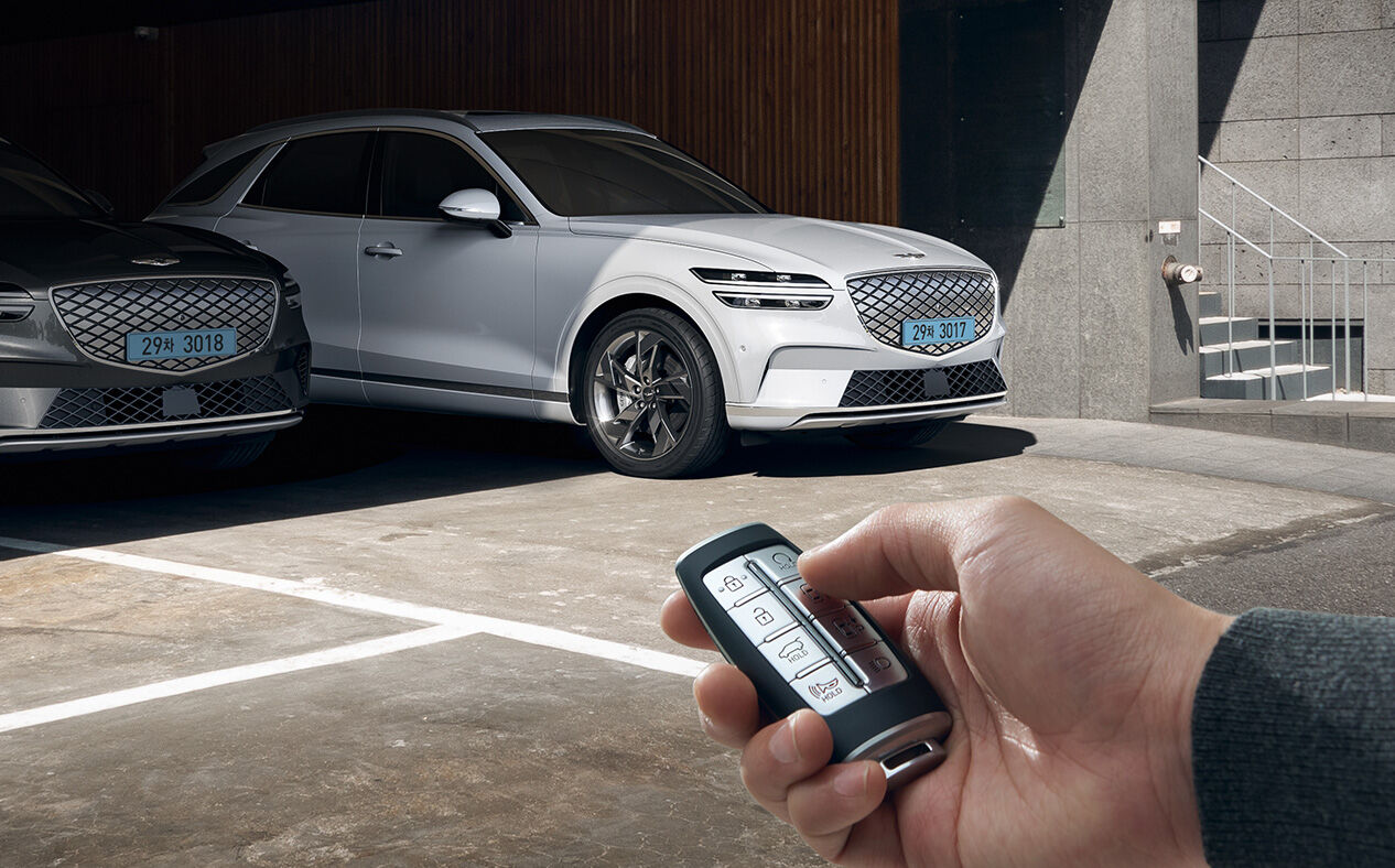 The Electrified GV70 is seen in the distance parked in a parking lot as a person holds a key and presses the Remote Smart Parking Assist button.