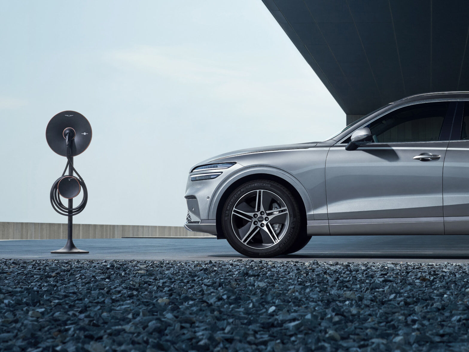 The Electrified GV70 is parked in front of a modern building, with a charger in front of the vehicle.