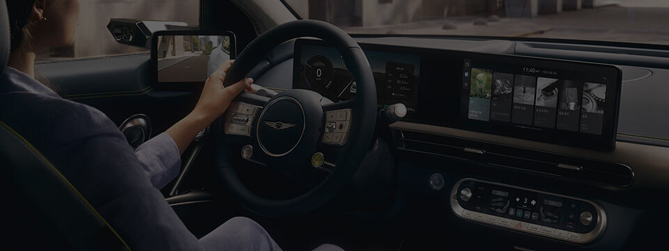 A woman sits in the driver's seat of the Genesis GV60, holds the steering wheel, and sees the spacious interior embodying Genesis' design philosophy.