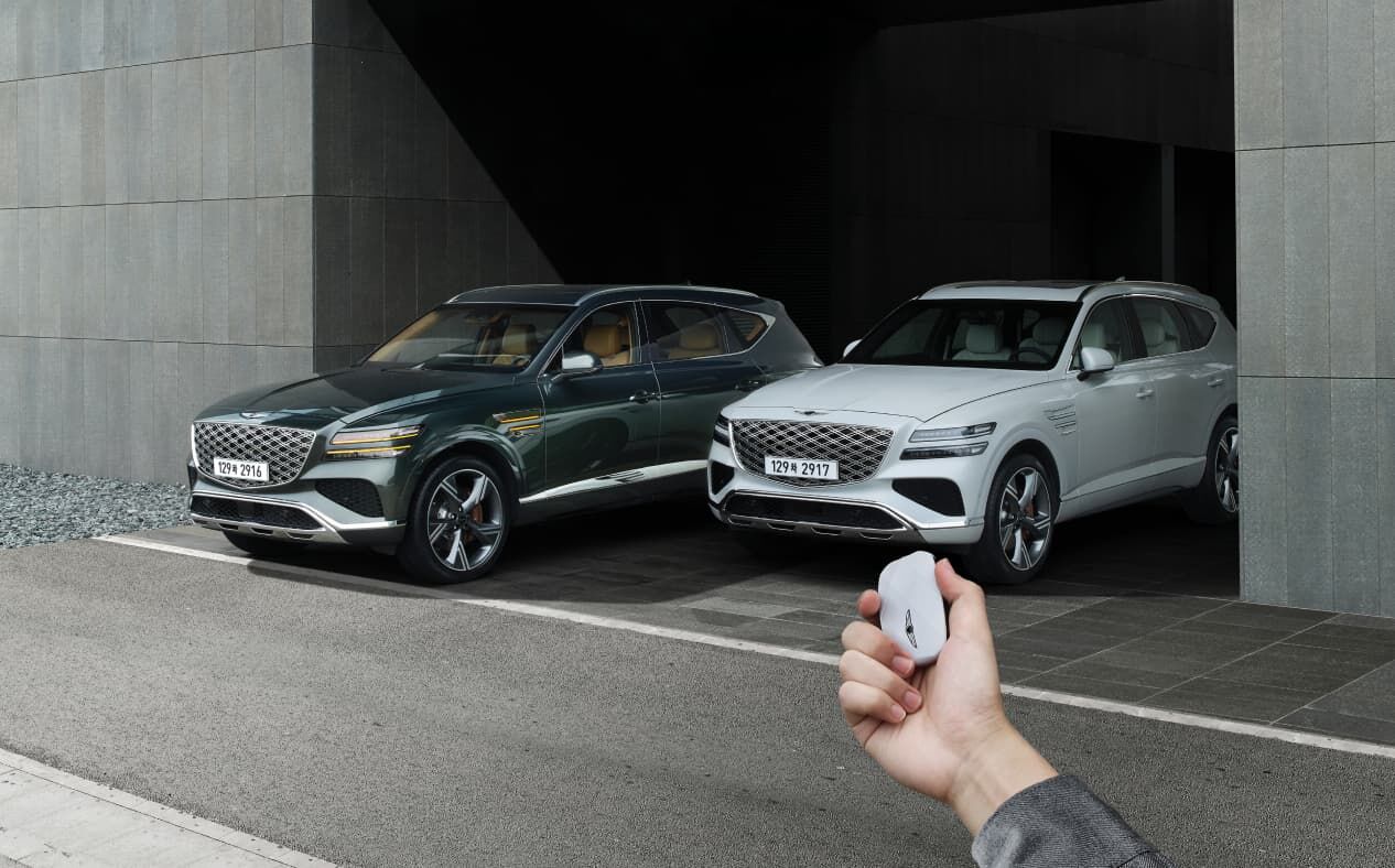A Vik Black GV80 and a Uyuni White GV80 are parked, and the driver is operating the smart key outside the vehicle.