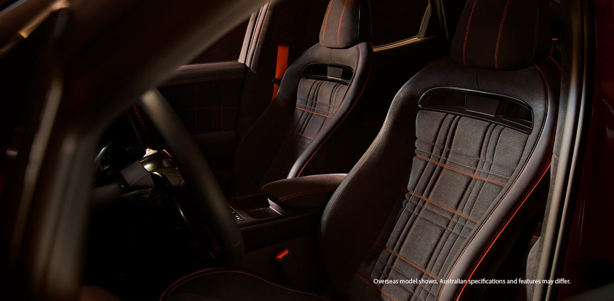 Close-up of Genesis GV60 Magma interior featuring sporty front bucket seats with perforated upholstery, integrated headrests, and orange contrast stitching, illuminated by warm ambient lighting that highlights the premium materials and performance-focused